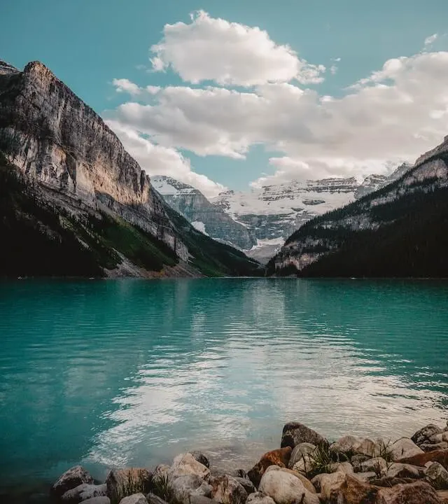 Lake Louise turquoise waters surrounded by mountains under a blue sky with white fluffy clouds.