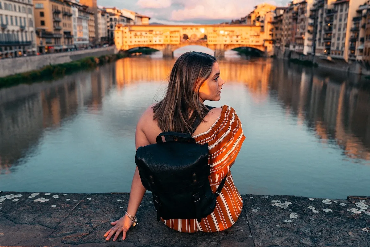 Travel Essentials for Women cover photo of a women sitting with her back to the camera wearing a black small backpack with the Ponte Veccio Bridge in Florence in the background
