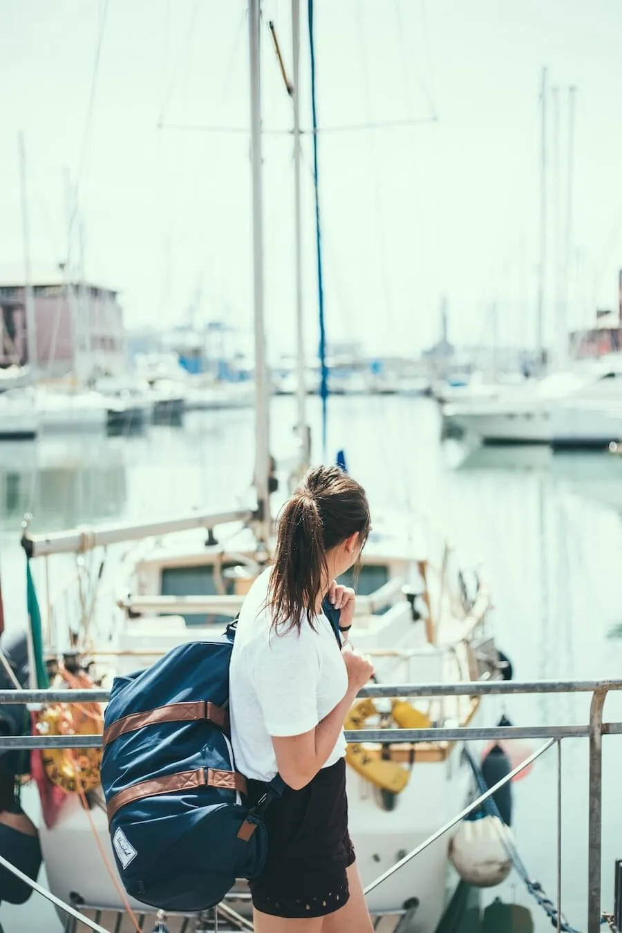 Women's Travel Essentials List image of a woman holding a large duffel bag over her shoulder while standing above a marina looking out at the water and the yachts below.