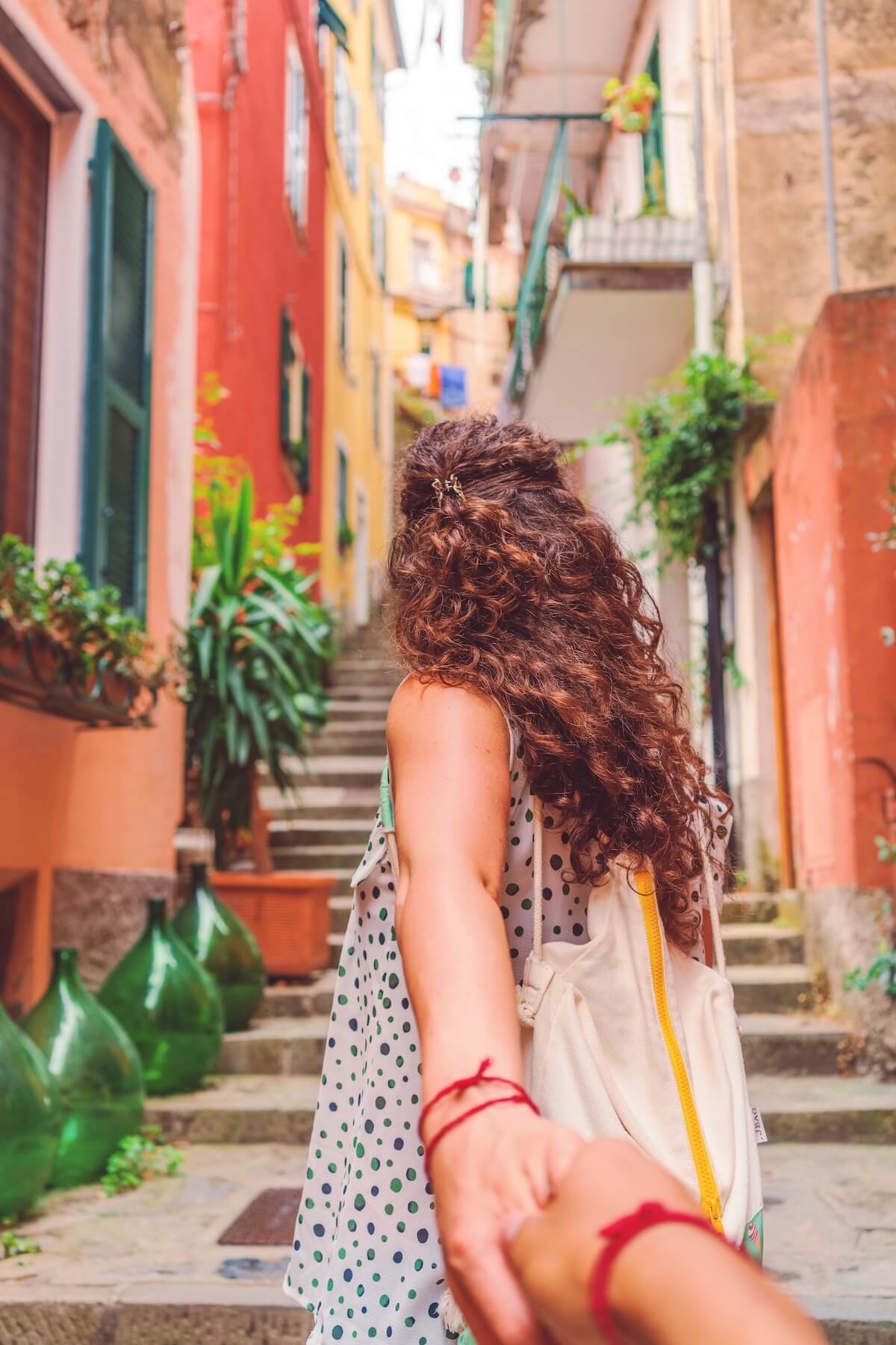 Europe Travel Essentials Checklist photo of a woman with long curly brown hair wearing a sundress leading an unknown person up a narrow colourful street in Europe