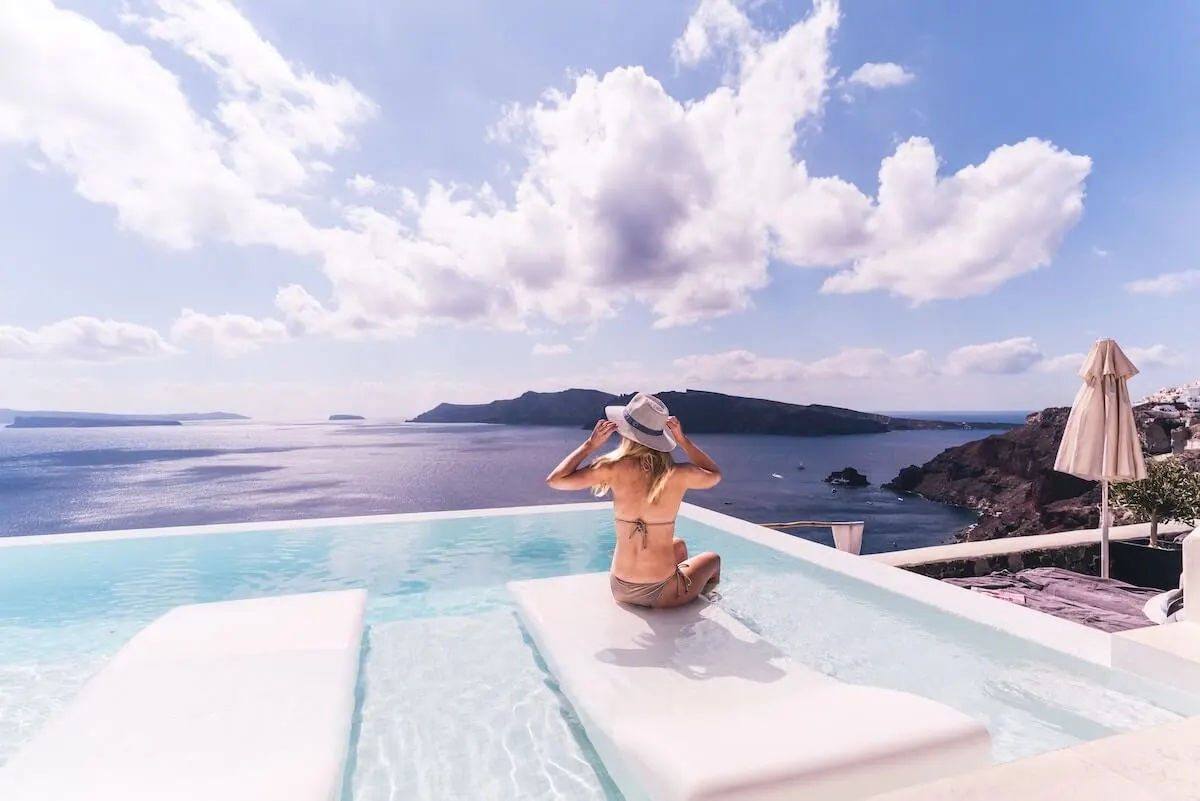 Travel Essentials for Europe cover photo of a woman in a bikini sitting in an infinity pool overlooking the ocean in Oia, Santorini