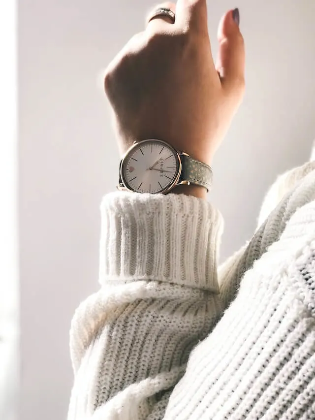 Arm of a woman wearing a white chunky jumper and a large wristwatch in front of a white wall