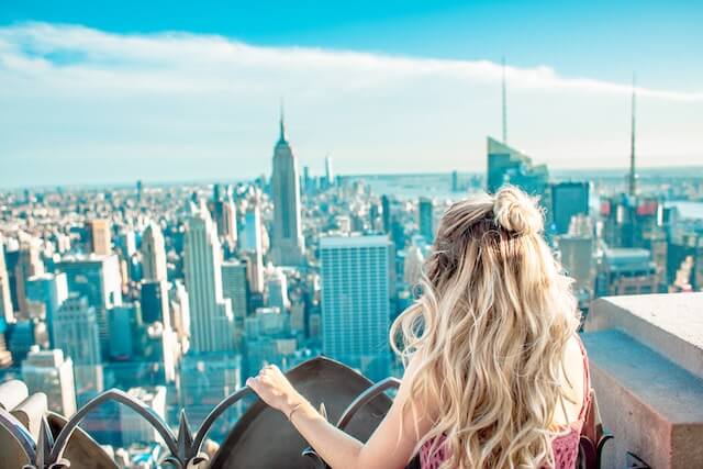 Blonde haired woman in a pink dress standing on the top of a building overlooking New York City