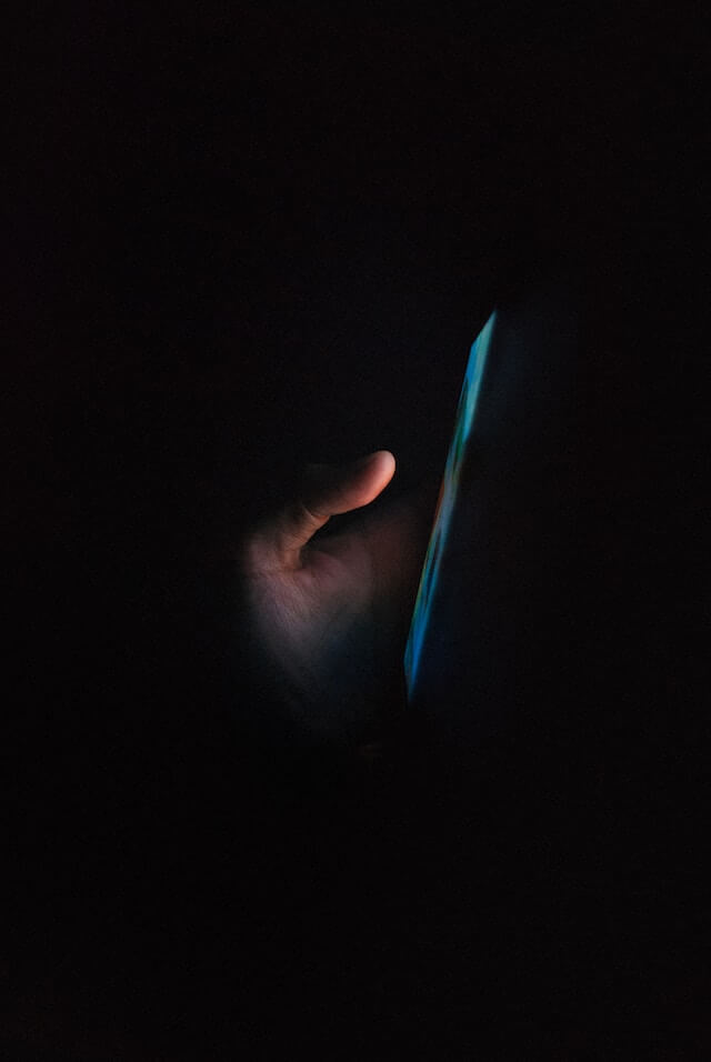 Hand holding a mobile phone, light from the screen illuminating the hand in an otherwise dark space