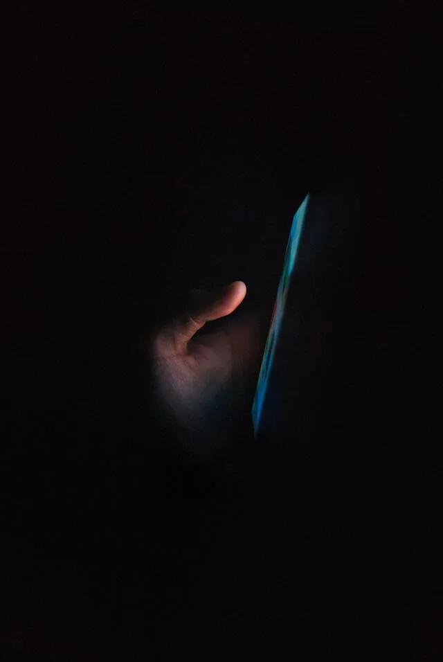 Hand holding a mobile phone, light from the screen illuminating the hand in an otherwise dark space