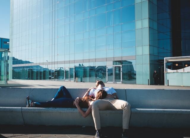 Man sat sleeping on a concrete bench, a woman lay on her back with her head resting on his leg, sleeping