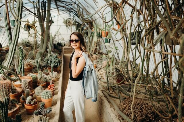 Woman wearing a black top with white pants and black sunglasses standing inside the greenhouse of Moorten Botanical Garden Palm Springs