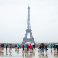 Rainy Day in Paris Things to Do cover photo of tourists standing on Trocadero Square in the rain, holding umbrellas looking at the Eiffel Tower, which looks misty in the distance