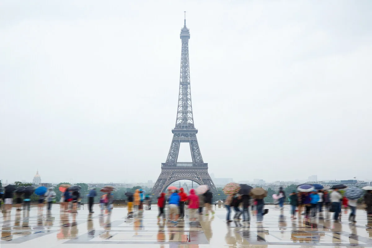 Rainy Day in Paris Things to Do cover photo of tourists standing on Trocadero Square in the rain, holding umbrellas looking at the Eiffel Tower, which looks misty in the distance